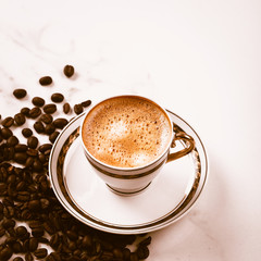 Cup of fresh americano or espresso coffee with golden foam froth on pile of brown raw coffee beans on white marble table background. Morning hot drink, coffee break, cope space