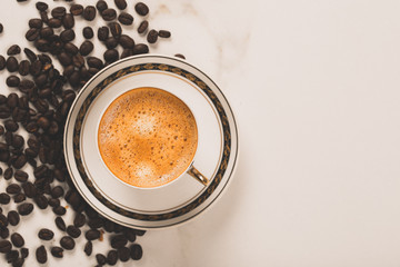 Cup of fresh americano or espresso coffee with golden foam froth on pile of brown raw coffee beans on white marble table background. Morning hot drink, coffee break, cope space