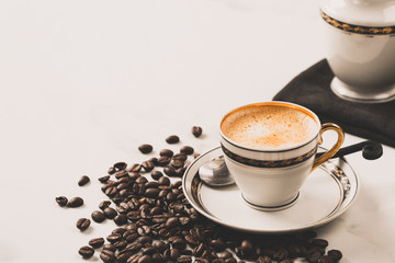 Cup of fresh americano or espresso coffee with golden foam froth on pile of brown raw coffee beans on white marble table background. Morning hot drink, coffee break, cope space