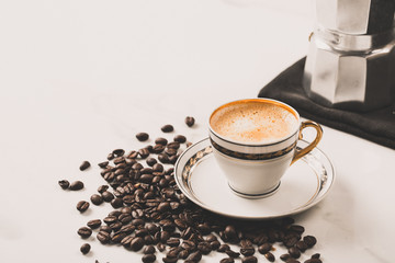 Cup of fresh americano or espresso coffee with golden foam froth on pile of brown raw coffee beans on white marble table background. Morning hot drink, coffee break, cope space