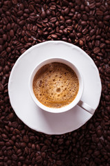 Cup of fresh americano or espresso coffee with golden foam froth on pile of brown raw coffee beans on white marble table background. Morning hot drink, coffee break, cope space