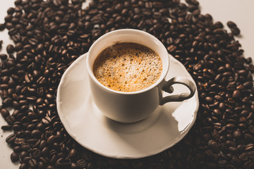 Cup of fresh americano or espresso coffee with golden foam froth on pile of brown raw coffee beans on white marble table background. Morning hot drink, coffee break, cope space