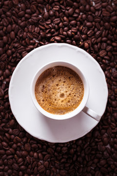 Cup of fresh americano or espresso coffee with golden foam froth on pile of brown raw coffee beans on white marble table background. Morning hot drink, coffee break, cope space © Chinara