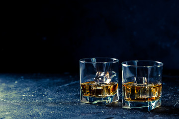 Two glasses of whiskey with ice cubes served on wooden board. Dark background, glossy golden color hard liquor on countertop with highlight. Night life bar relaxation concept