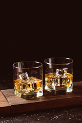 Two glasses of whiskey with ice cubes served on wooden board. Dark background, glossy golden color hard liquor on countertop with highlight. Night life bar relaxation concept