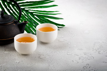 Asian style tea ceremony, black iron kettle teapot with white bowl cups with palm leaf on light background. Healthy hot detox drink, copy space