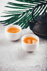 Asian style tea ceremony, black iron kettle teapot with white bowl cups with palm leaf on light background. Healthy hot detox drink, copy space