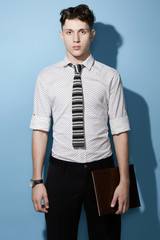 Fototapeta premium Medium full shot of a young dark haired man in a black trousers and white printed shirt with a knit tie with black, gray and white stripes. The man with a black watch is holding a leather folder. 