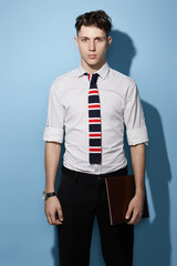 Fototapeta premium Medium full shot of a young dark haired man in a black trousers and white printed shirt with a deep blue knit tie with red and white stripes. The man with a black watch is holding a leather folder. 