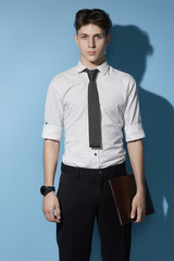 Fototapeta premium Medium full shot of a young dark haired man in a black trousers and white printed shirt with a gray knit tie with little gray dots. The man with a black watch is holding a leather folder. 