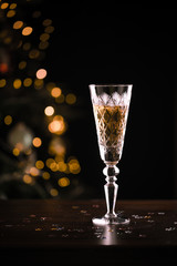 Glass of sparkling champagne for Christmas or midnight New Year night celebration, festive arrangement, wooden table, sparkling garland lights, dinnerware, copy space with bokeh background