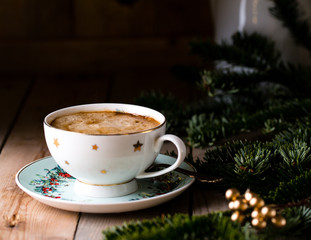 Cup of coffee or eggnog on vintage rustic wooden table with bokeh wreath background, Christmas morning concept decoration, beautiful festive cup with stars, green pine branch