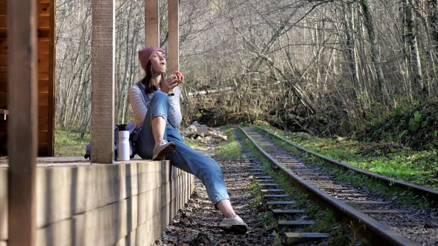 Young backpacker woman in denim overall is sitting on country train station alone and eating apple