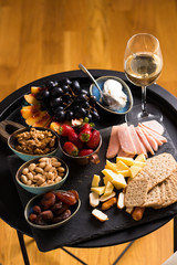 Cheese and fruits plate with glass of white wine on round tray, colorful and delicious charcuterie with snacks and wineglass for romantic evening, wine tasting, selective focus.