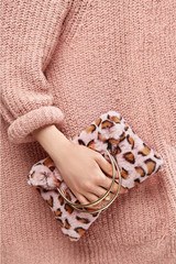 Close-up cropped shot of a girl, wearing pale pink knitted sweater. She's holding pale pink leopard print faux fur bag with metallic semicircular bag handles. There are two golden rings on her fingers