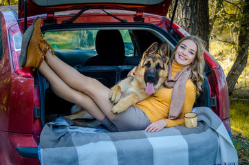 A happy Caucasian girl lies in the trunk of a car with a German shepherd dog breed and smiles, a cheerful playful teen puppy looks at the camera. Friendship of man and animal, travel, camping