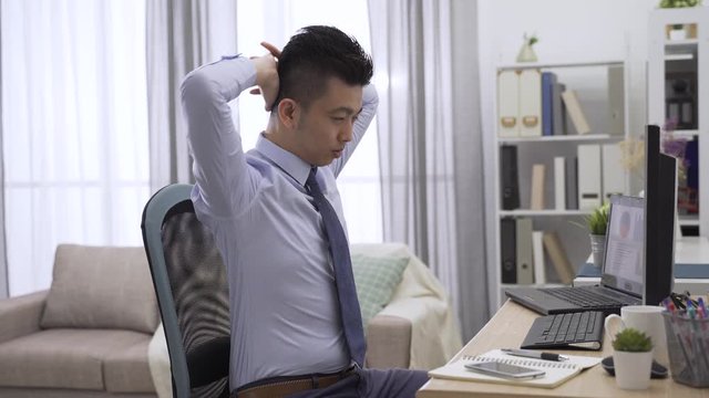 asian man freelance worker working in home workplace stretching back while long time typing computer. male businessman doing easy office exercises to relieve muscle tension from sedentary work