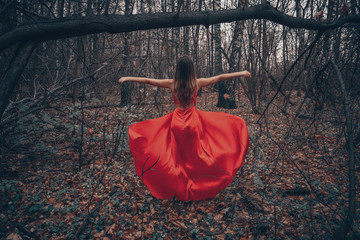 Young beautiful woman in the long flying red dress is walking along foggy mysterious forest