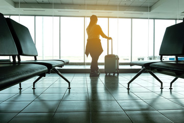 Silhouettes of Female passenger standing and Look through the window to the outside of the building with luggage, While waiting for the delayed plane concept.
