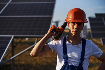 Using cordless screwdriver. Male worker in blue uniform outdoors with solar batteries at sunny day