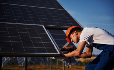 Using cordless screwdriver. Male worker in blue uniform outdoors with solar batteries at sunny day