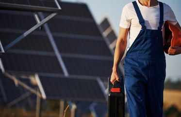 Holds case with equipment in hand. Male worker in blue uniform outdoors with solar batteries at...
