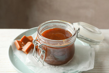 Glass jar with salted caramel and candies on wooden background, space for text