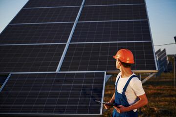 Holds notepad in hand. Male worker in blue uniform outdoors with solar batteries at sunny day