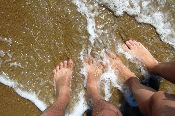 Fototapeta premium The legs of a man and a woman stand on the sandy shore of the sea and they are slightly doused with water