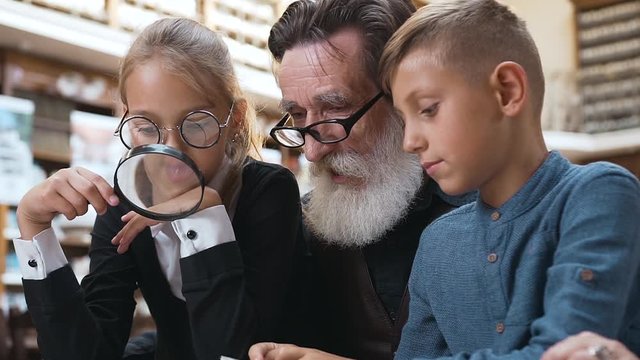 Appealing blond grandchildren sitting at the library table with their respected eldery grandpa and reading book using magnifying glass