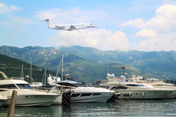 Fototapeta na wymiar Private jet business plane flies low over yachts and boats in gulf of background of mountains with white clouds