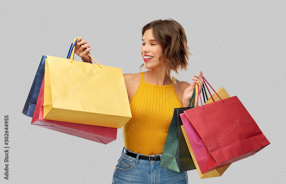 Sticker sale and people concept - happy smiling young woman in mustard yellow top and jeans with shopping bags over grey background