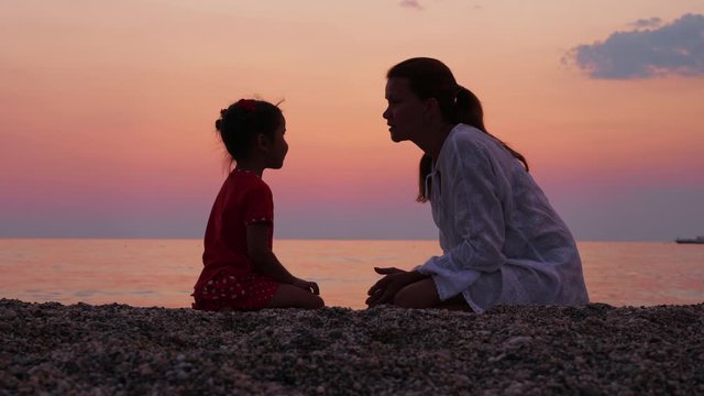 Mother talk to her little daughter by the sea or ocean shore at sunset. Mom speak to baby, they have fun and kiss while sitting at the beach at sundown. Trust and motherhood concept.