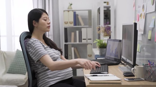 Young asian chinese pregnant business woman using laptop computer at bright home office. motherhood lady freelancer worker sitting at workplace typing on keyboard indoors. future mom touching belly