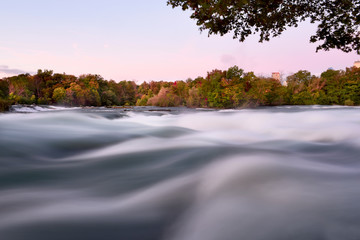 View of the Niagara River in the autumn early morning. Bright colorful trees and a rapid stream of water running to the cliff of Niagara Falls. USA. Long exposure. Blurred running water.