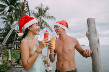 Young couple with cocktails in Christmas caps on the ocean on a tropical island.