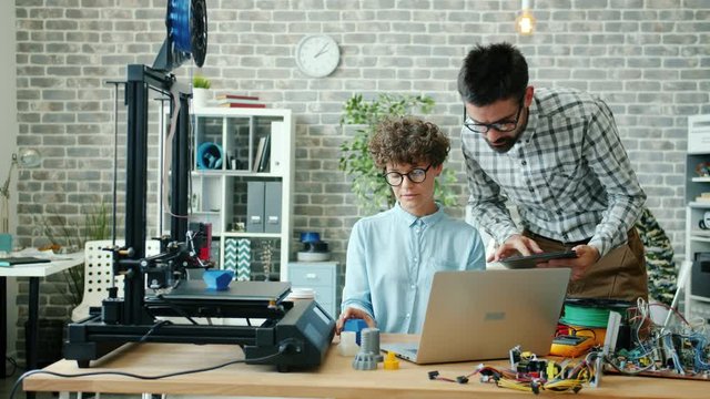 Man and woman are using tablet, laptop and 3d printer in office talking working together with modern technology. People, teamwork and business concept.