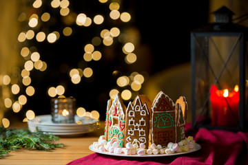 Little gingerbread houses with glaze standing on table with red tablecloth and decorations, candles and lanterns. Lights of Christmas tree on background.  Powdered sugar falling like snow flakes