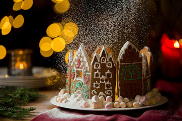 Little gingerbread houses with glaze standing on table with red tablecloth and decorations, candles and lanterns. Lights of Christmas tree on background.  Powdered sugar falling like snow flakes