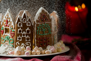 Little gingerbread houses with glaze standing on table with red tablecloth and decorations, candles and lanterns. Lights of Christmas tree on background.  Powdered sugar falling like snow flakes
