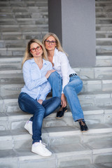 A beautiful blonde with glasses and her mature mother are sitting on the stairs outdoor. A well-groomed elderly woman hugs her adult daughter tightly. Love and family values. Mothers Day.