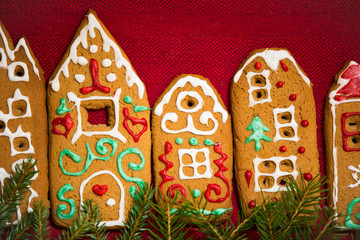 Christmas background. Red table cloth with town of cute gingerbread houses decorated with icing, Christmas lights, glitter and fir tree branches. Holiday mood. 