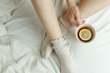 Fototapeta na wymiar Cozy flatlay of woman's legs in warm white stockings in bed holding cup of lemon tea, selective focus