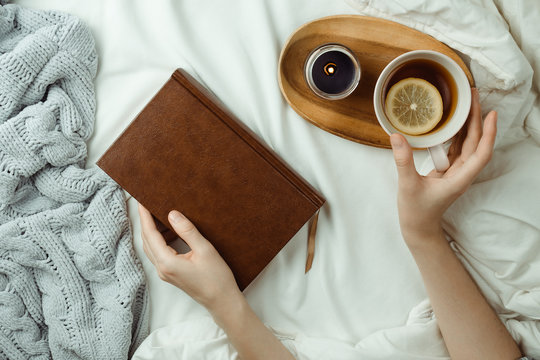 Cozy flatlay of woman's hand holding lemon tea and book in bed with sheets, blanket, knitted sweater and candle