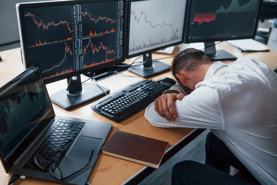 Tired businessman sleeping on the workplace by leaning on the table with multiple screens on it. Stock information on displays