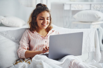 Cute girl lying on the bed with laptop in the bedroom at her weekend time
