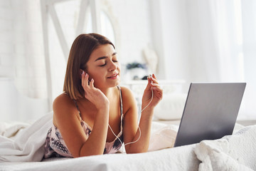 Listening to the music by using headphones. Cute girl lying on the bed with laptop in the bedroom...