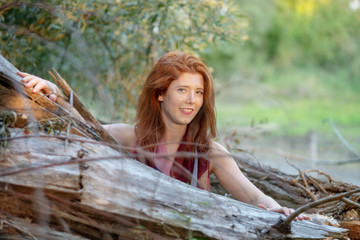 beautiful young red-haired girl looks nicely hidden behind a fallen gray tree trunk.