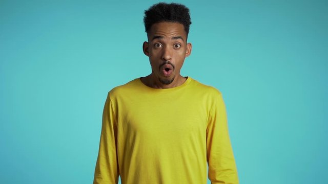 Amazed mixed race man shocked, saying WOW. Handsome african american guy with afro hair surprised to camera over blue background.