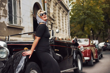 Fototapeta na wymiar Blonde woman in sunglasses and in black dress leaning on old vintage classic car